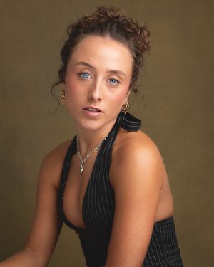 Portrait of a woman in a black pinstripe top against a brown backdrop, wearing a necklace and styled with curly hair.