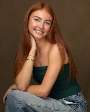 A woman with long red hair poses in a dark green top and jeans against a brown studio backdrop.