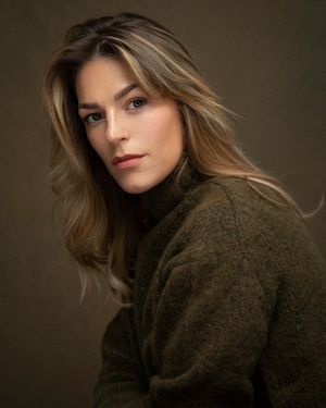 Portrait of a person in a brown turtleneck sweater with long wavy hair against a dark moody background.
