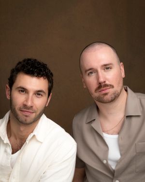 Two men in casual attire pose for a studio portrait against a brown backdrop.