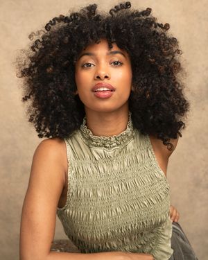 Model wearing a sage green smocked turtleneck top poses with natural curly hair against a neutral background.