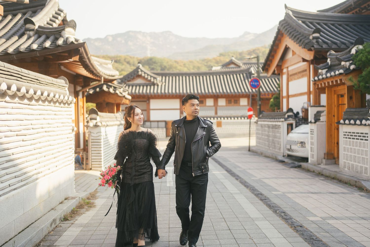 A couple holding hands while walking through traditional Korean architecture with tiled roofs and wooden buildings.