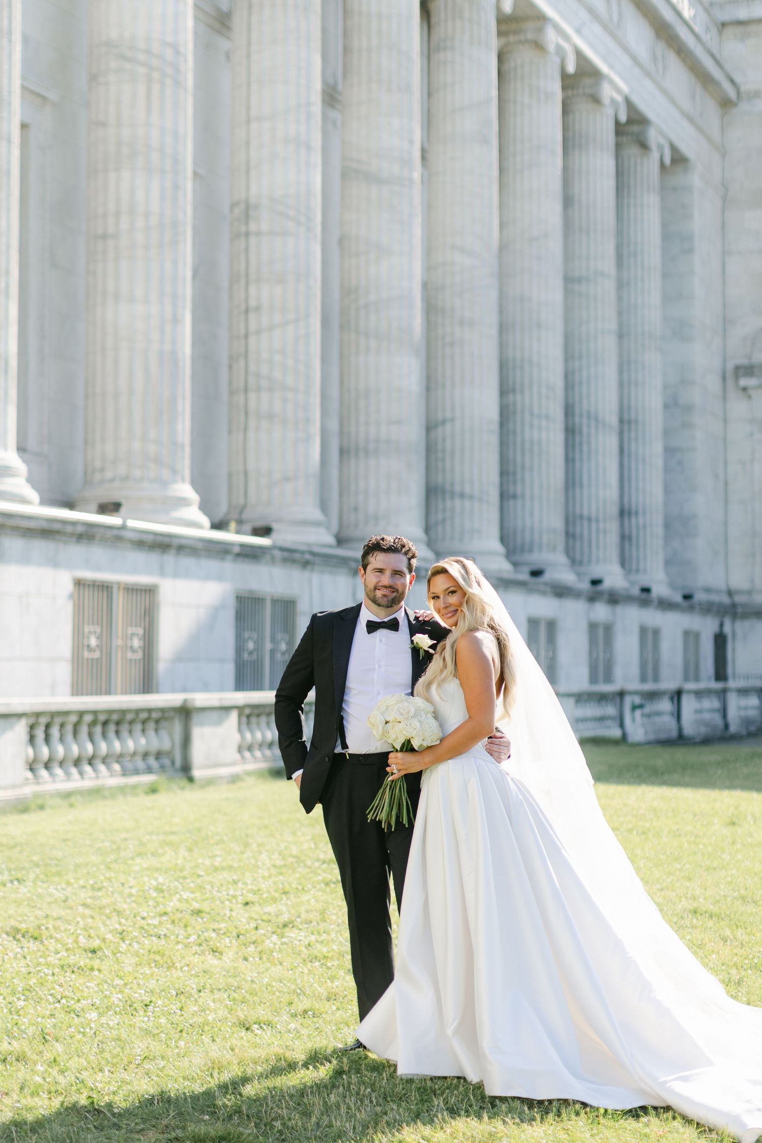 A bride in a flowing white dress twirls gracefully in front of a grand columned building on a sunny day.