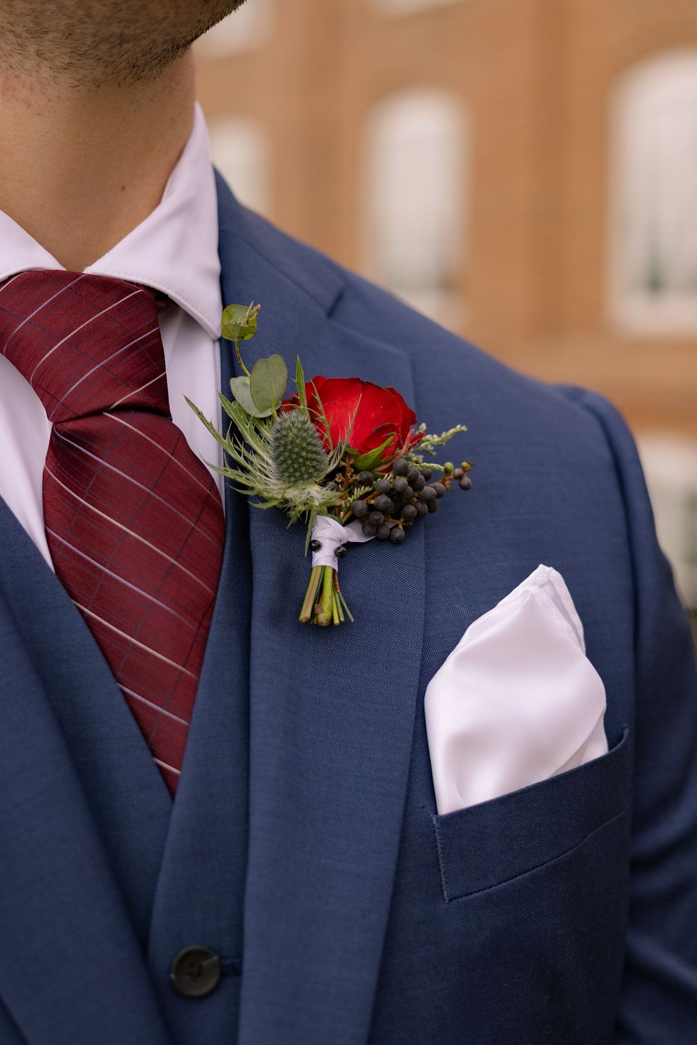 Close-up of navy blue suit jacket with burgundy tie and red rose boutonniere showing elegant wedding attire details.