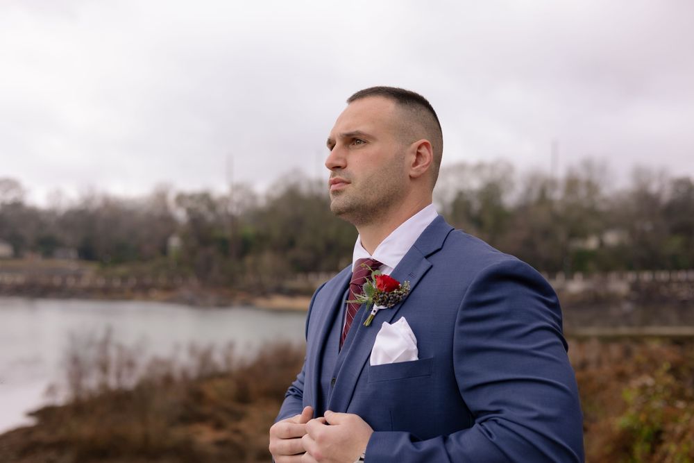 A person in a navy blue suit and red tie stands near a waterfront on an overcast day.