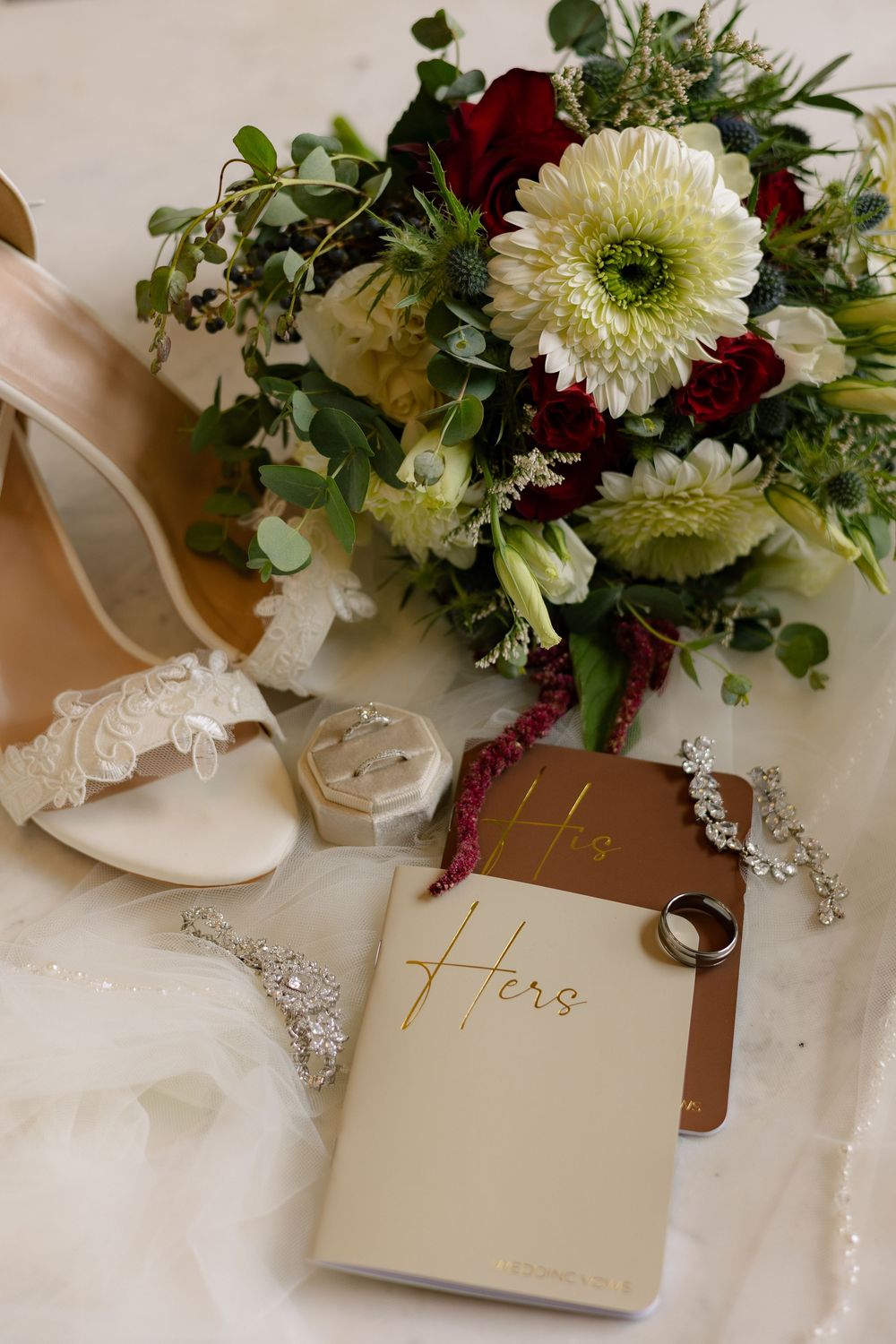 Wedding accessories including white shoes, floral bouquet with red and white flowers, and a vows book arranged elegantly.