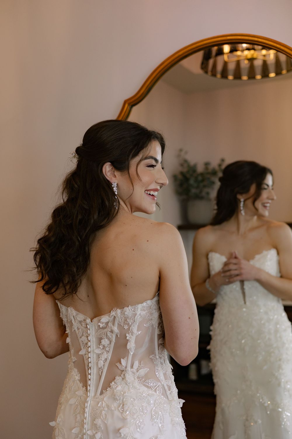 A bride in a detailed lace corset wedding dress smiles while looking in a mirror.