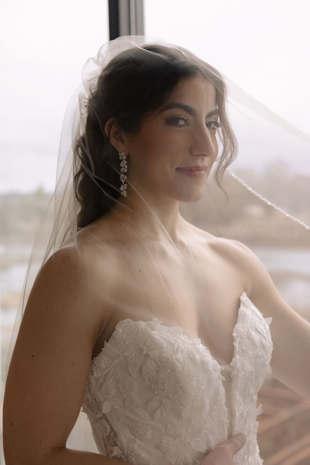 A bride wearing a strapless lace gown and veil poses by a window in soft natural light.