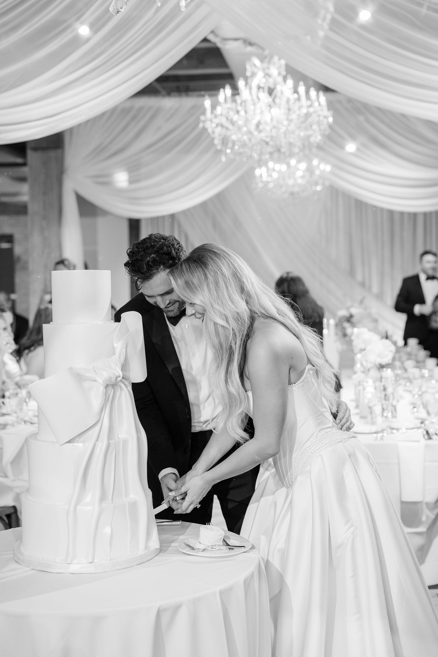 Elegant wedding reception setup with white draping, crystal chandeliers and romantic lighting in a rustic venue.