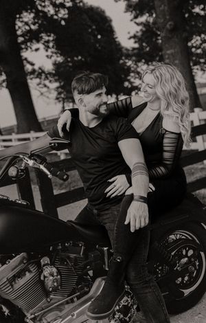 A romantic black and white photo of a couple sitting on a motorcycle in a park setting.