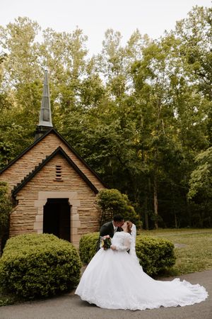 Bride and groom kissing in front of a charming stone chapel surrounded by lush green trees on their wedding day.