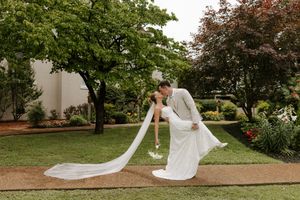 Bride and groom share a romantic dip kiss in a lush garden, with her long veil trailing gracefully on the grass.