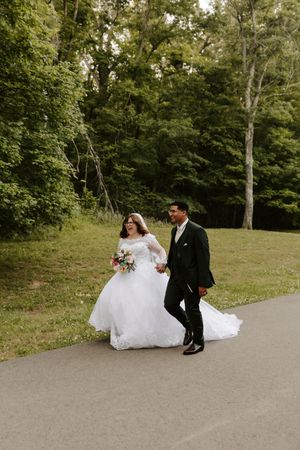 Bride and groom walking hand in hand on a path surrounded by lush green trees on their wedding day.