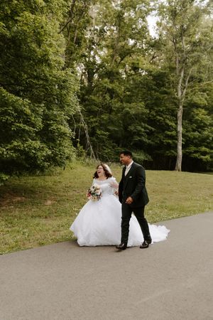 Bride in white ballgown and groom in dark suit walk together on a path surrounded by lush green trees.