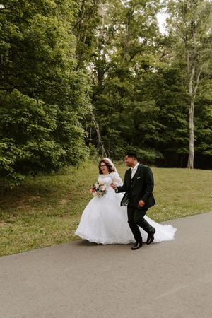 Bride in white gown and groom in dark suit walking together on a path surrounded by lush green trees.