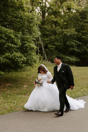 Bride and groom walk hand-in-hand on a path surrounded by lush green trees on their wedding day.