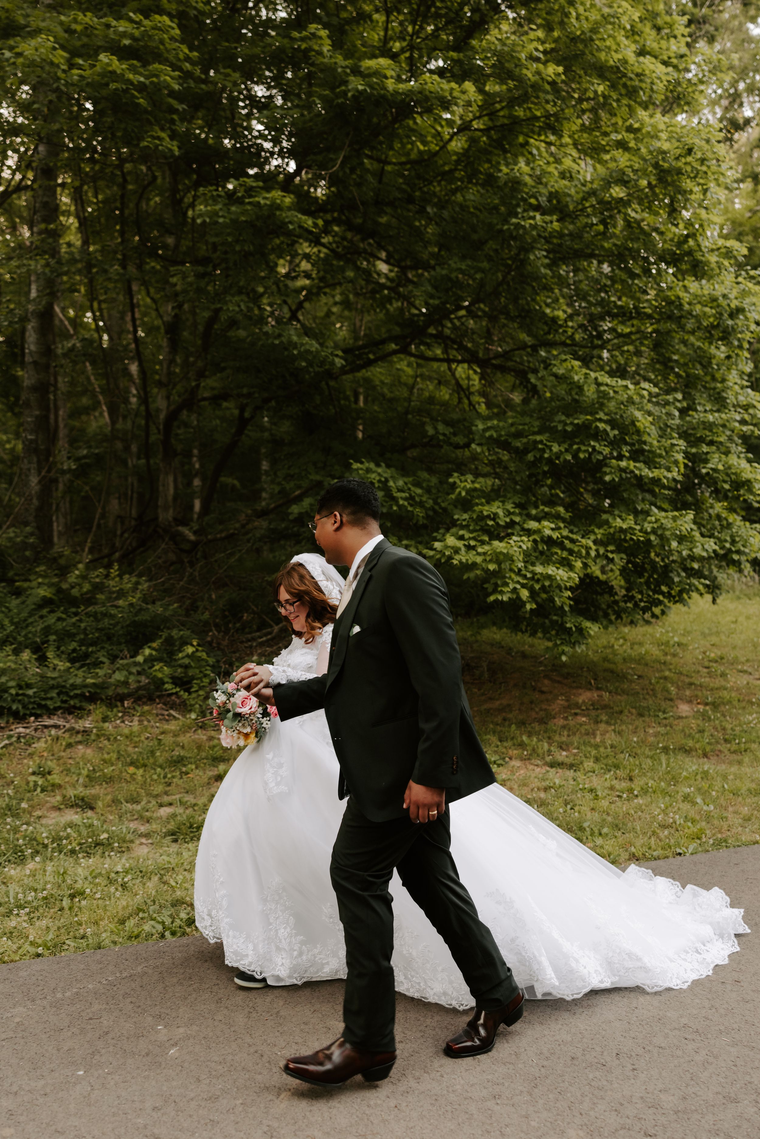 A bride and groom walk hand in hand on a path surrounded by lush green trees on their wedding day.