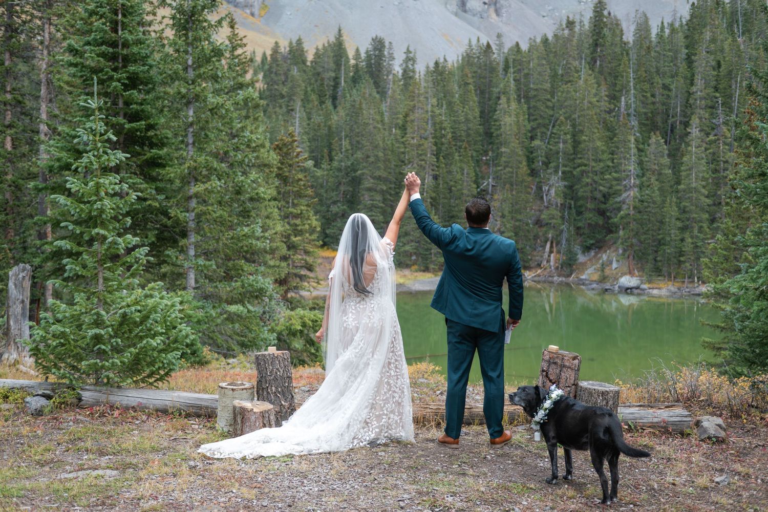 A bride and groom are holding hands together up in the air with their dog by mountain lake surrounded by pine trees at an outdoor wedding.