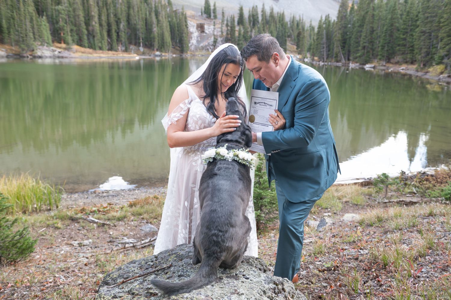 A bride and groom show their black dog their marriage license after a lakeside elopement ceremony in the mountains near Telluride.