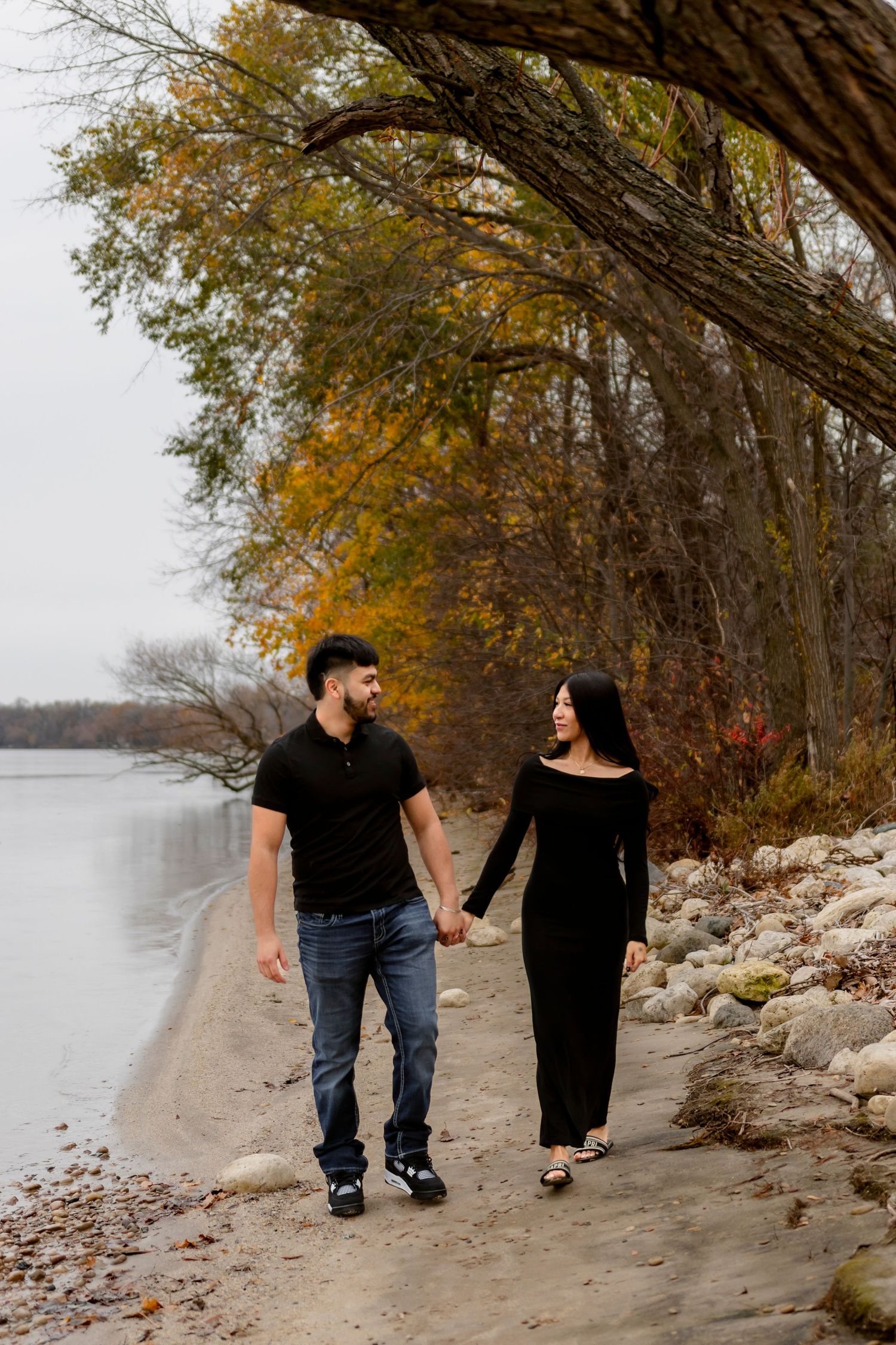 A couple walking hand in hand along a rocky beach shoreline during autumn with golden trees in the background.