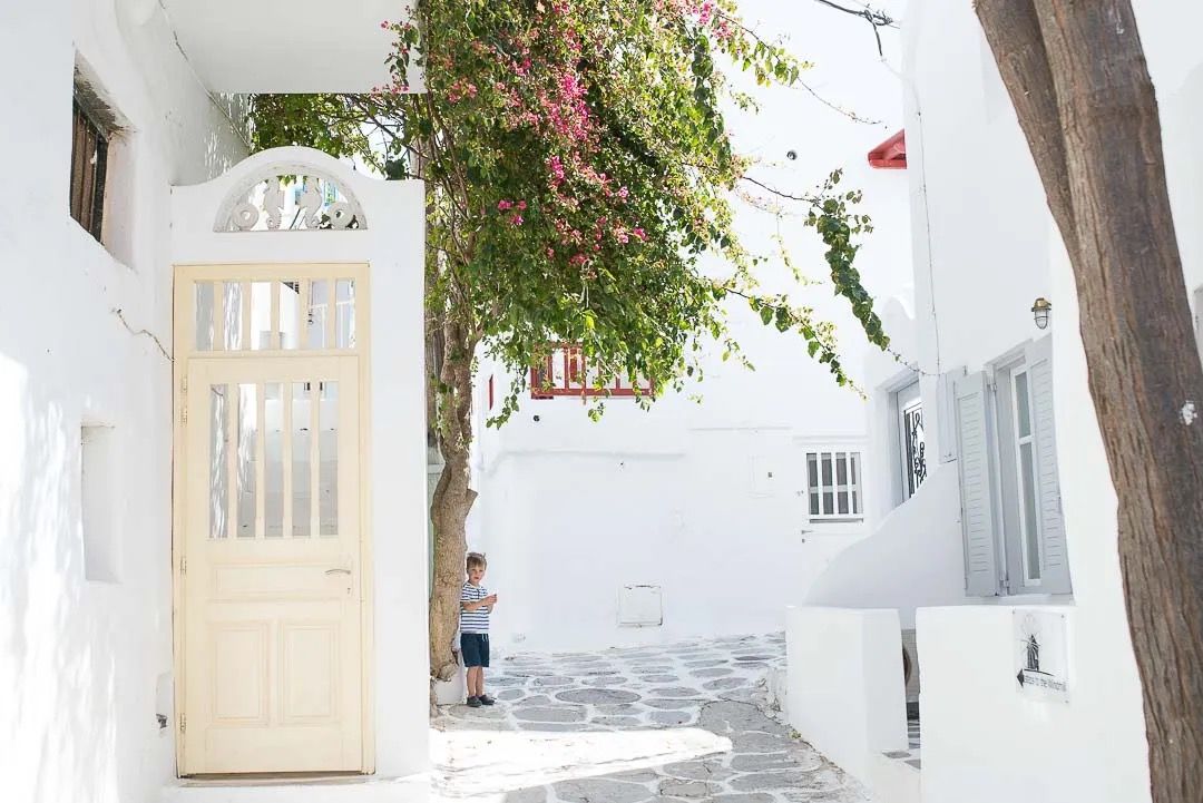 A charming white-walled Mediterranean alley with pink bougainvillea flowers cascading from above and wooden doors along the path.