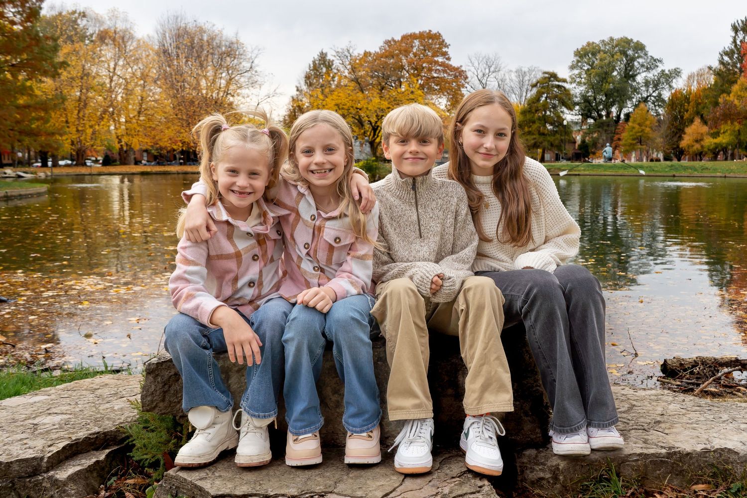 Family members sit together on a stone wall near a lake during autumn with colorful fall foliage in the background.