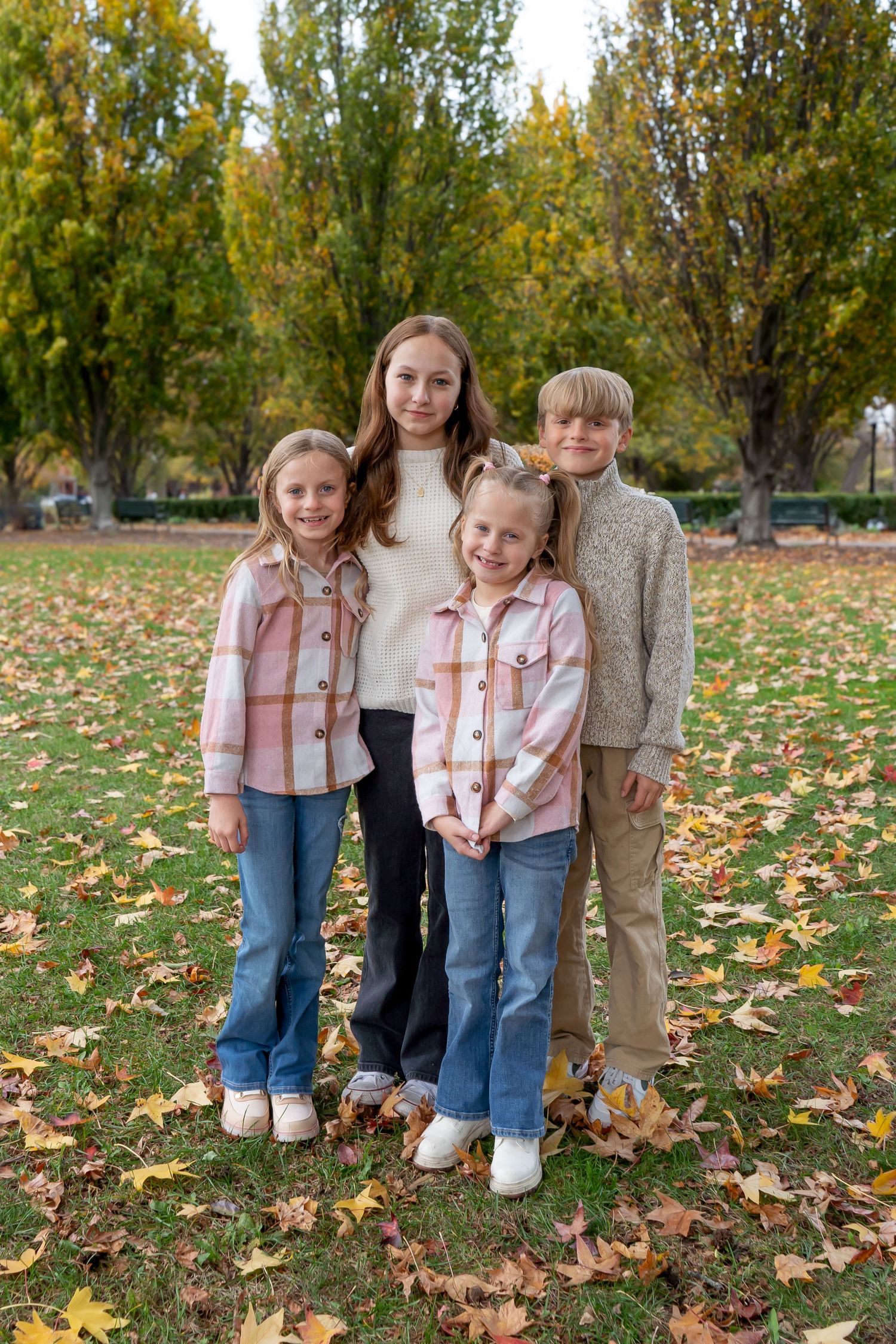 Three children stand together on fallen autumn leaves in a park setting with trees in the background.