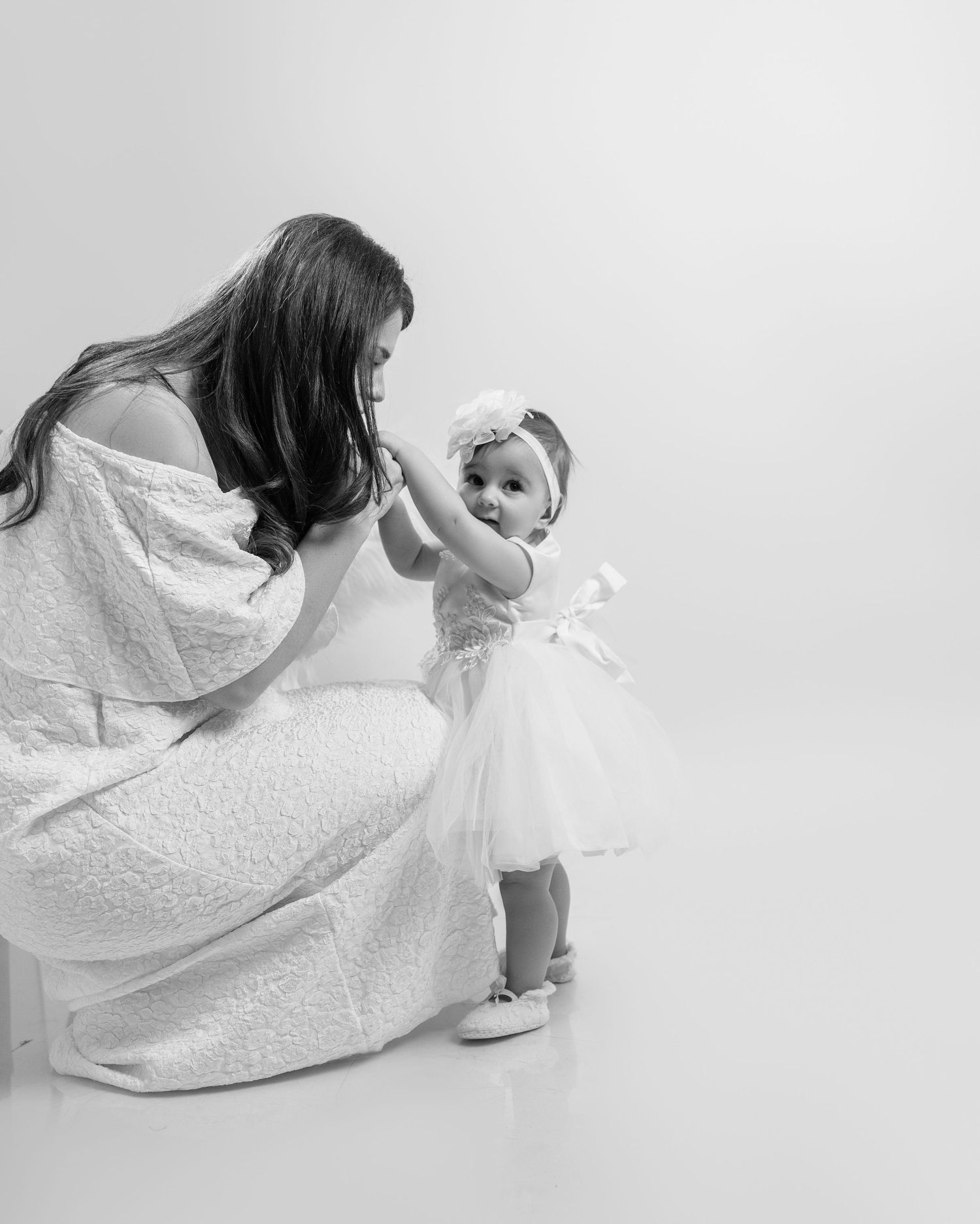 Mother in white dress interacting with baby girl in tutu dress in black and white studio photo.