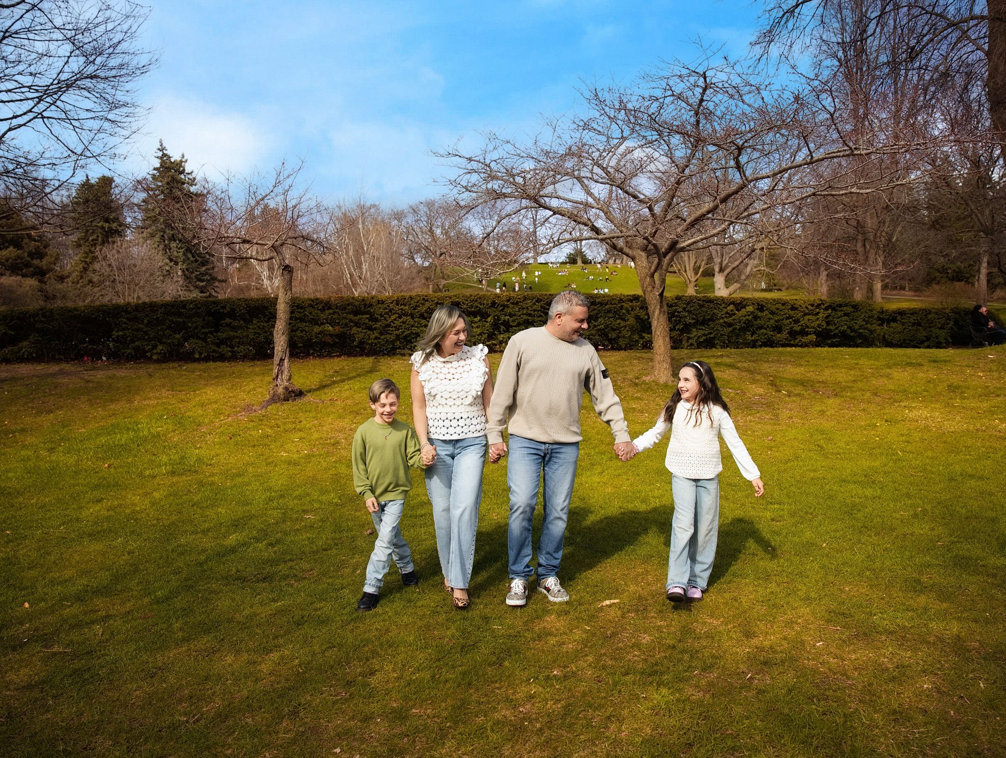 Happy family of four holding hands and walking on green grass in a park on a sunny day.