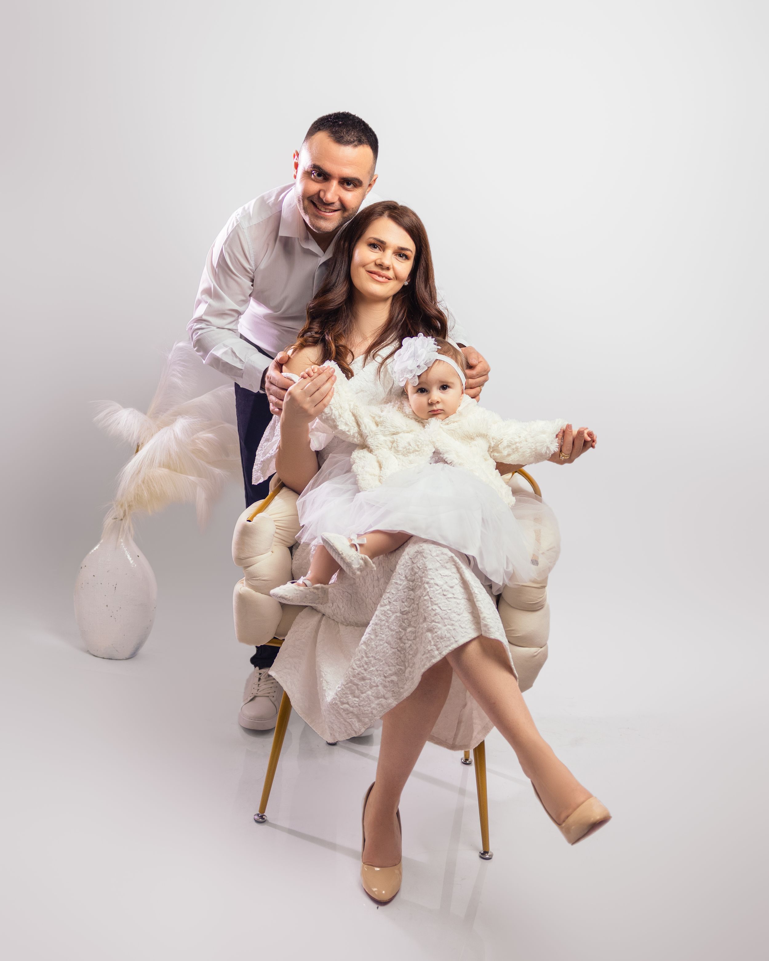 Family portrait with parents and baby in white outfits seated in a studio chair.