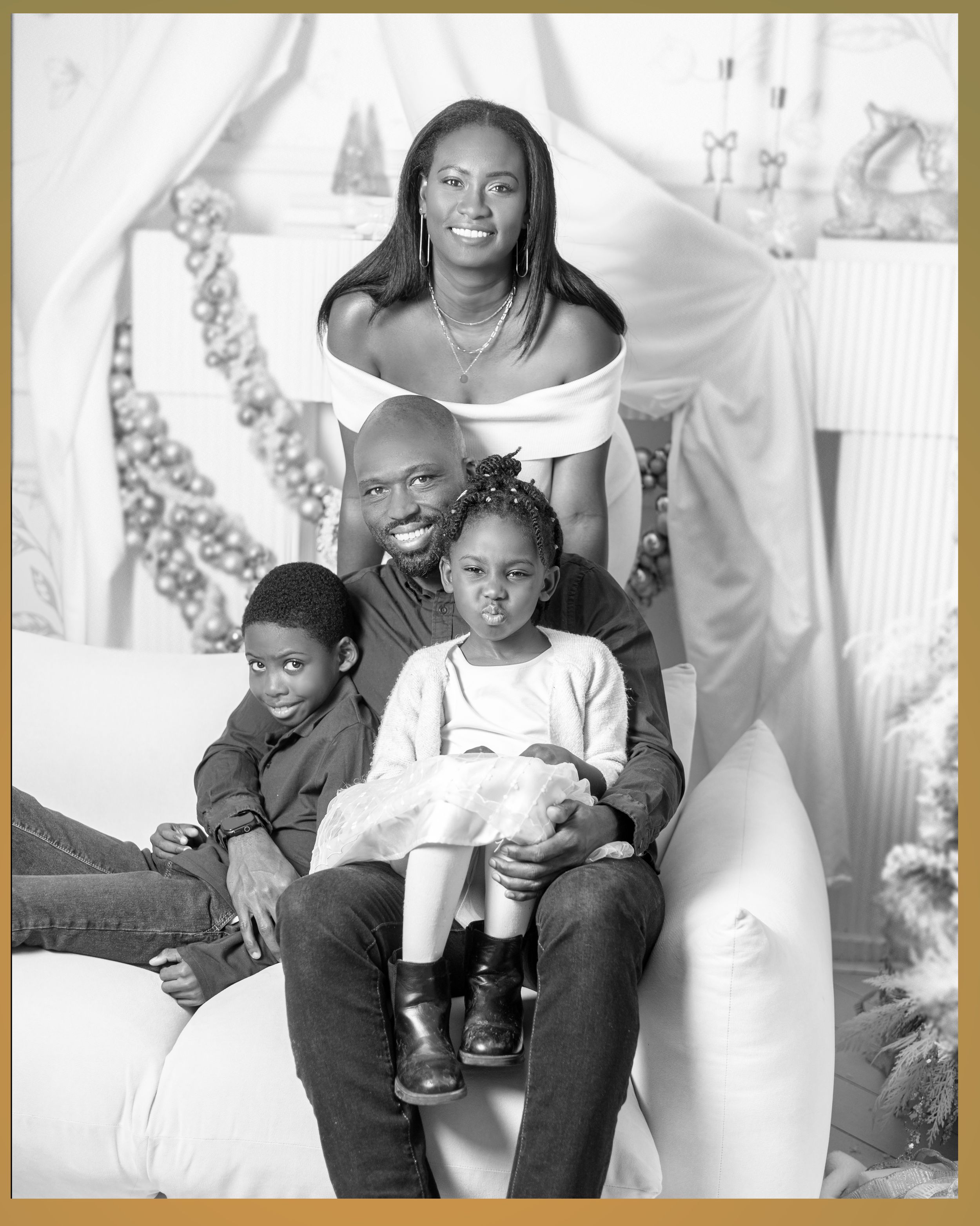 Black family of four poses together in black and white Christmas portrait on white sofa.