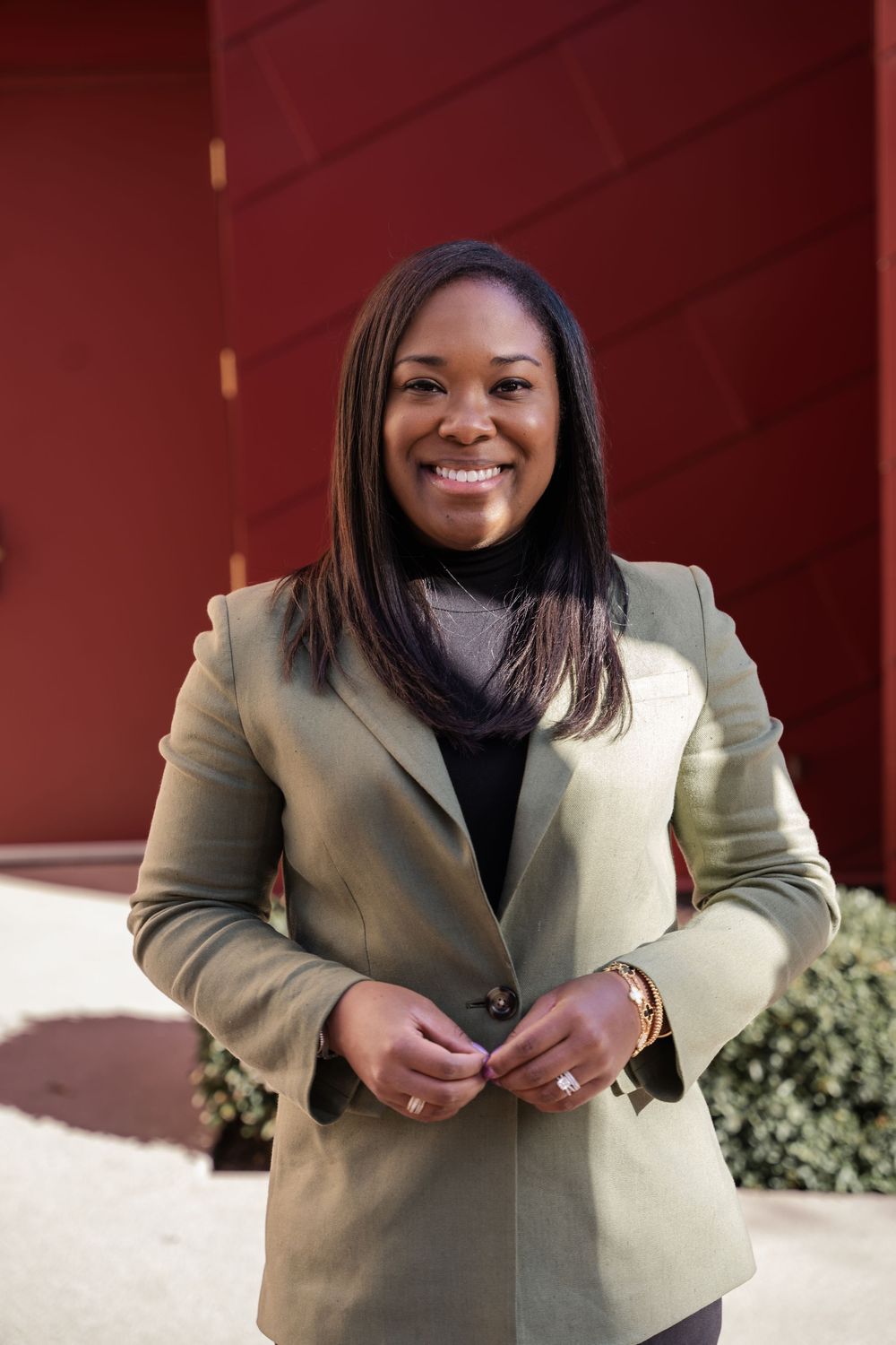 Professional portrait of someone wearing a beige blazer and black turtleneck against a red wall background.