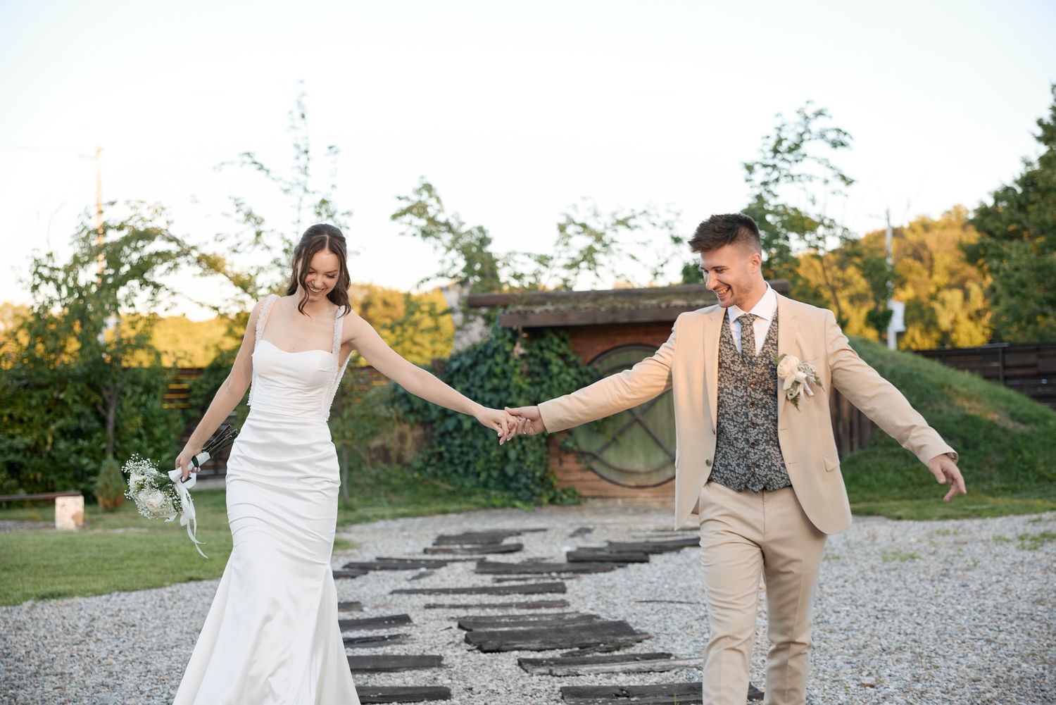 A couple in wedding attire holds hands while walking on a gravel path in a scenic garden setting.