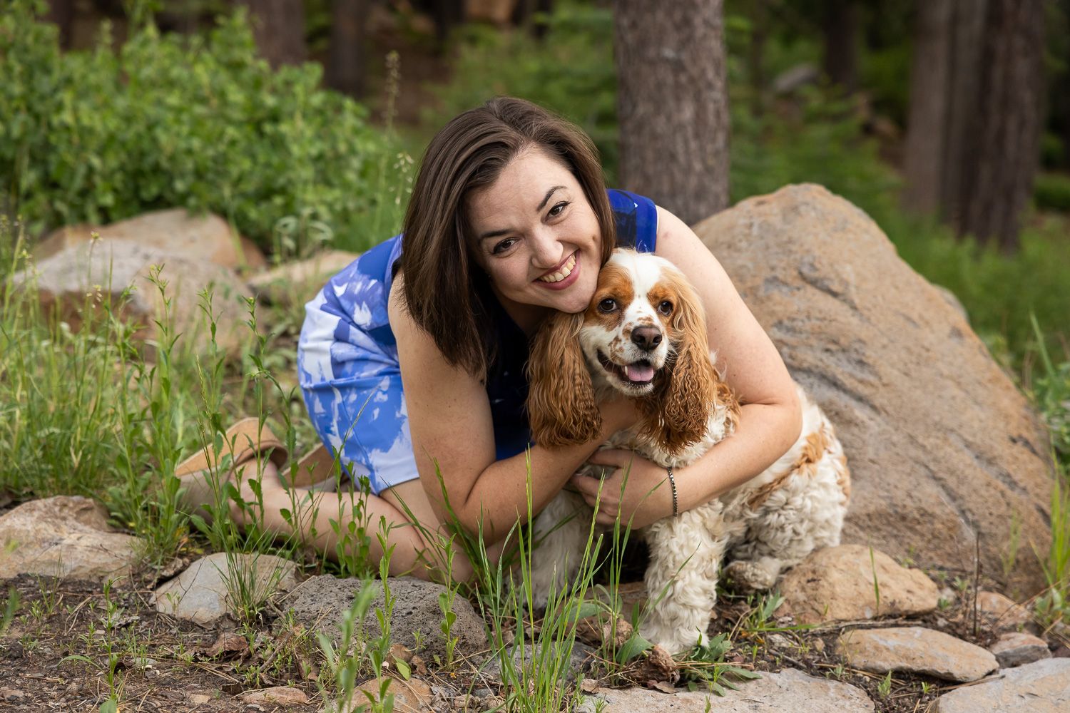 A person in a blue dress hugs her Cocker Spaniel dog while sitting on rocks in a wooded outdoor setting.