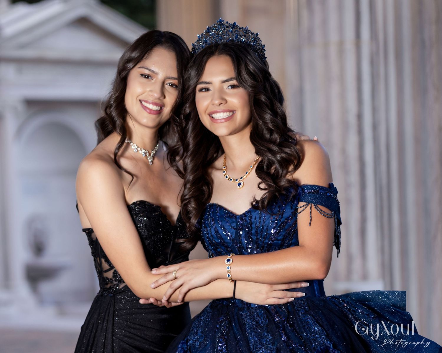 Two people in formal attire pose together on stone steps, one wearing black and one in a sparkling navy blue dress.