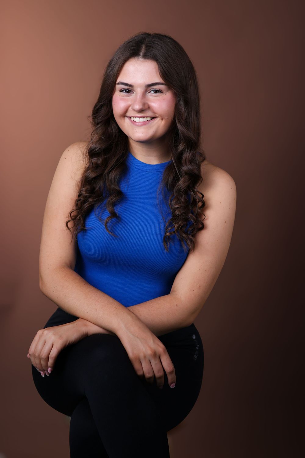 Studio portrait against brown background featuring a bright blue sleeveless top and long dark curly hair.