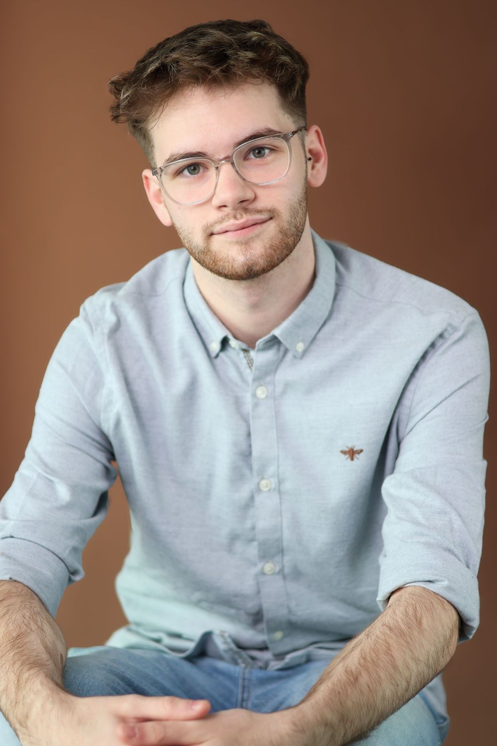 Professional portrait of someone wearing glasses and a light blue button-down shirt against a brown backdrop.