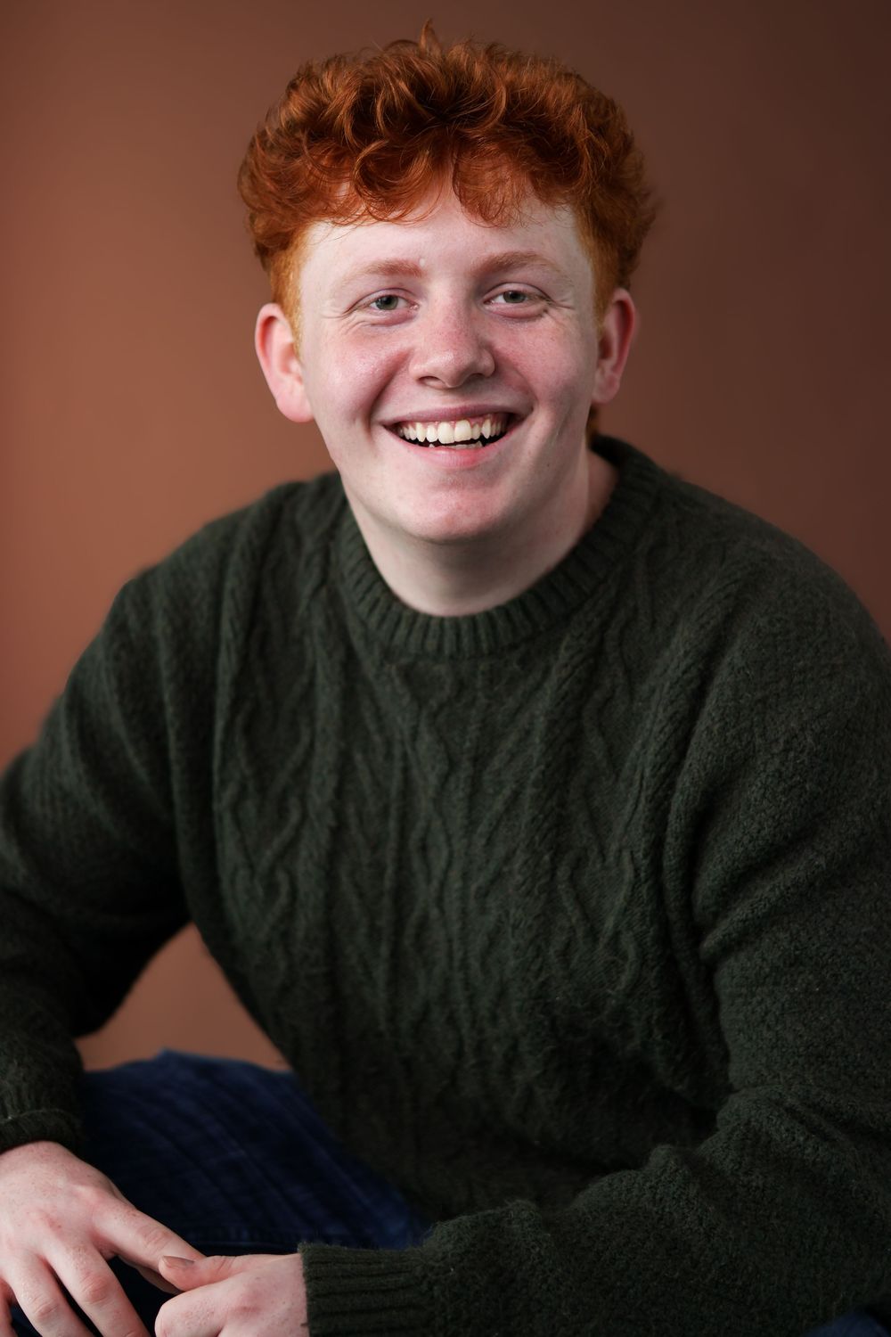 A smiling person with red hair wearing a dark green sweater poses against a brown background.