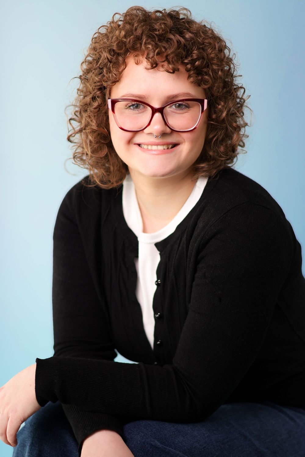 Professional headshot against blue backdrop showing person with curly hair, glasses, and black blazer smiling warmly.