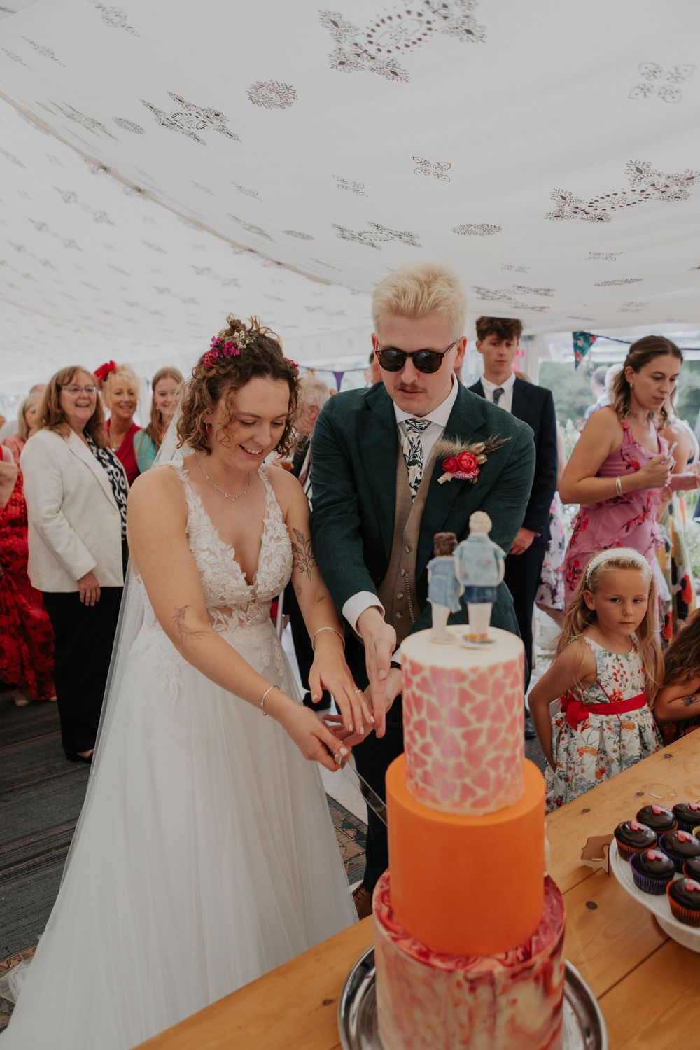 Bride and groom cutting a vibrant orange and pink tiered wedding cake under a decorated marquee tent.