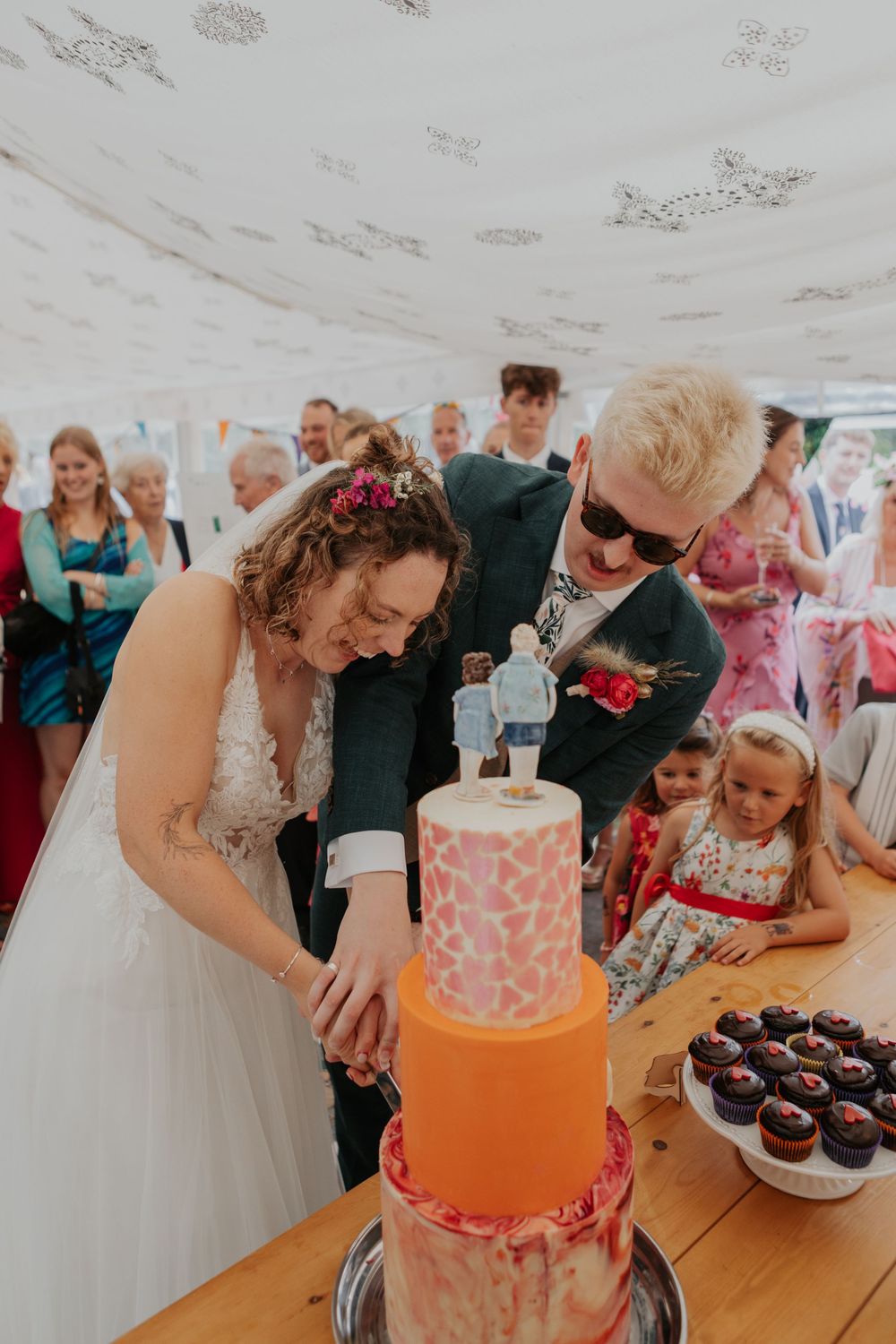Smiling bride and groom cutting their colorful orange wedding cake together surrounded by wedding guests in a marquee.