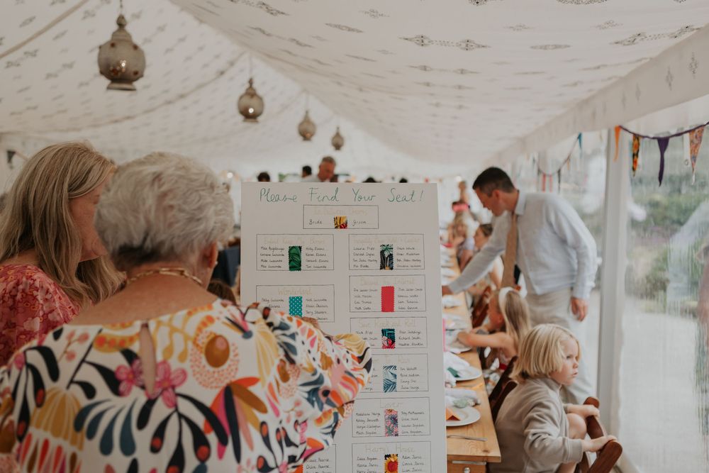 Guests browse a seating chart display inside a decorated wedding marquee with colorful bunting.