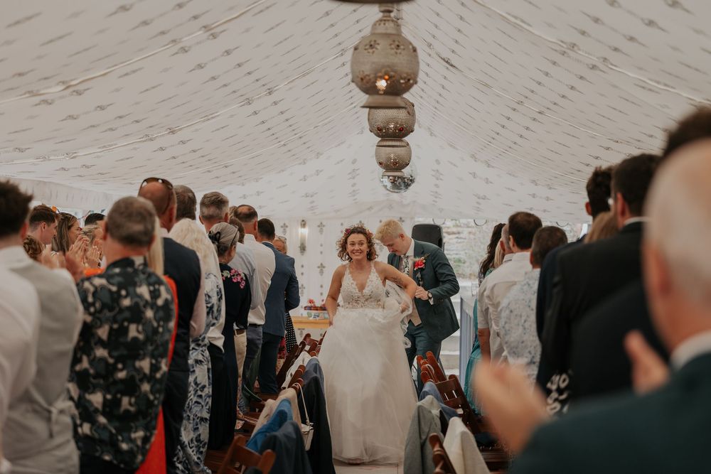 Radiant bride walking through a crowded marquee reception tent lined with cheering wedding guests.
