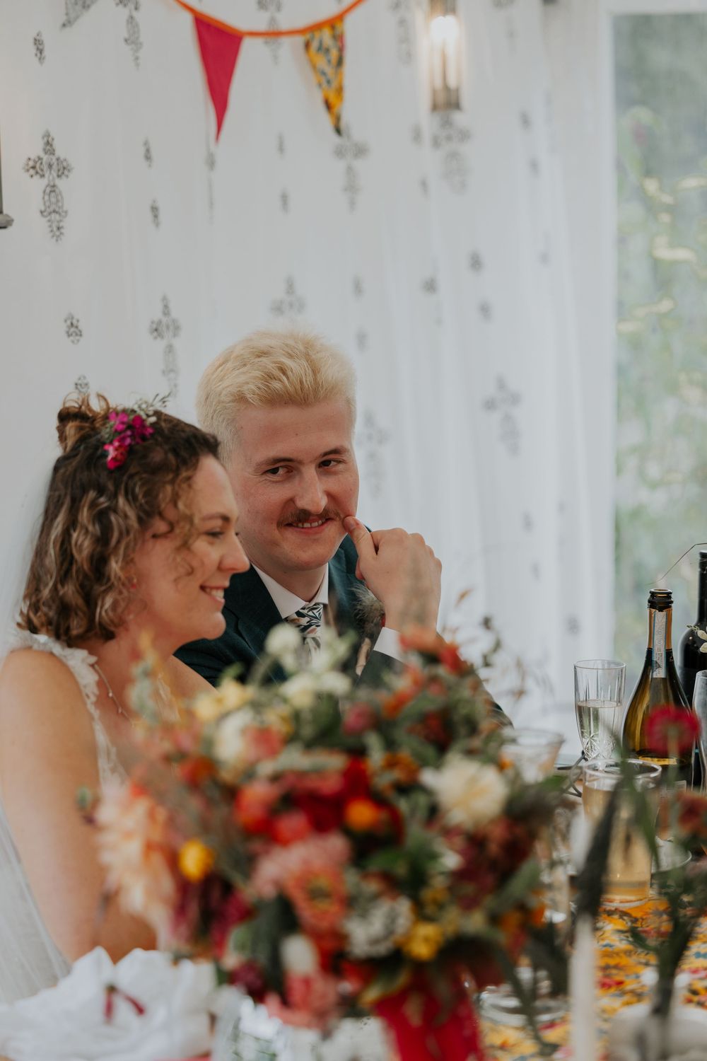 Smiling bride and groom sit together at the wedding reception table with floral centerpiece.