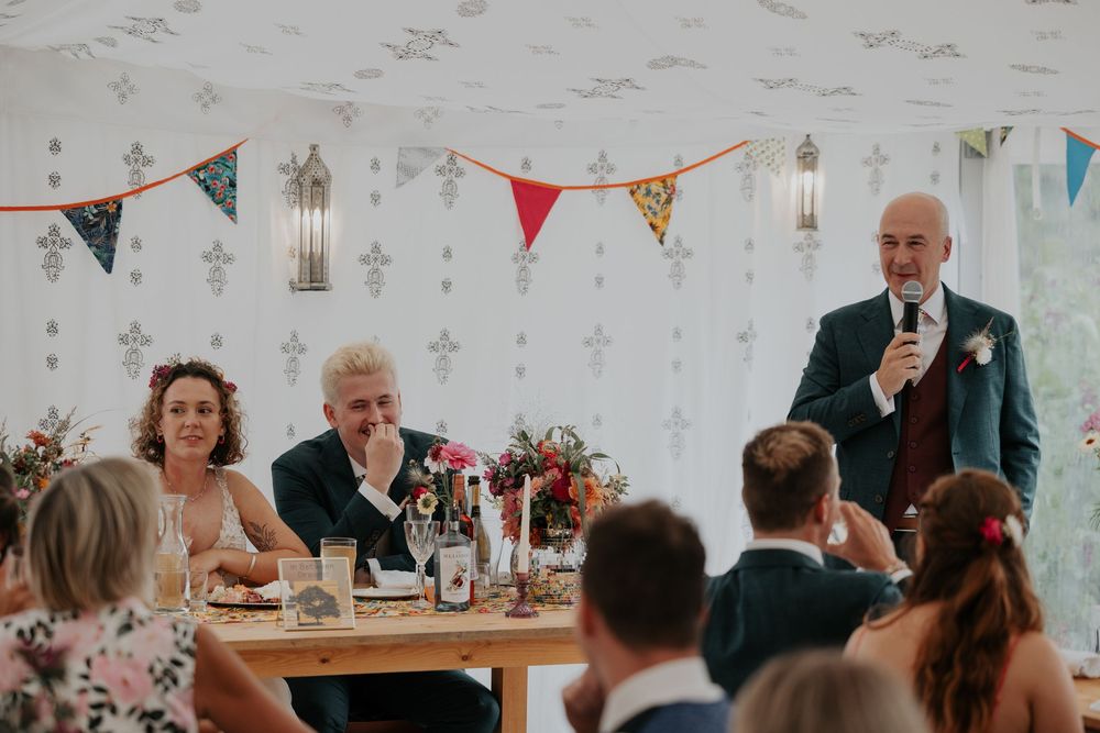 Man delivers a wedding speech at a decorated reception table while guests listen attentively.