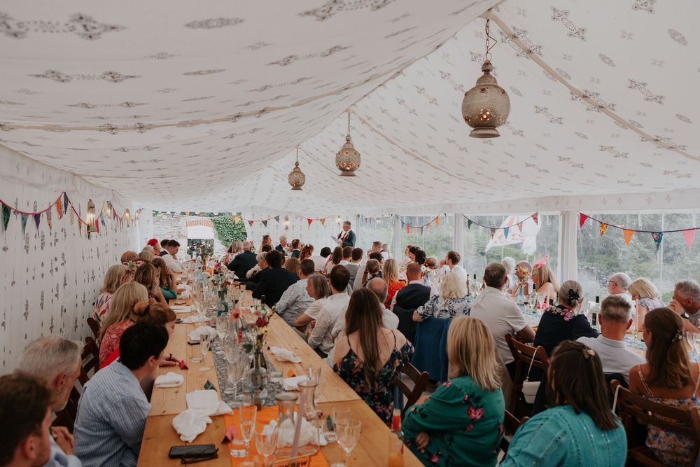 Wedding guests seated along long banquet tables inside a beautifully decorated marquee with bunting.