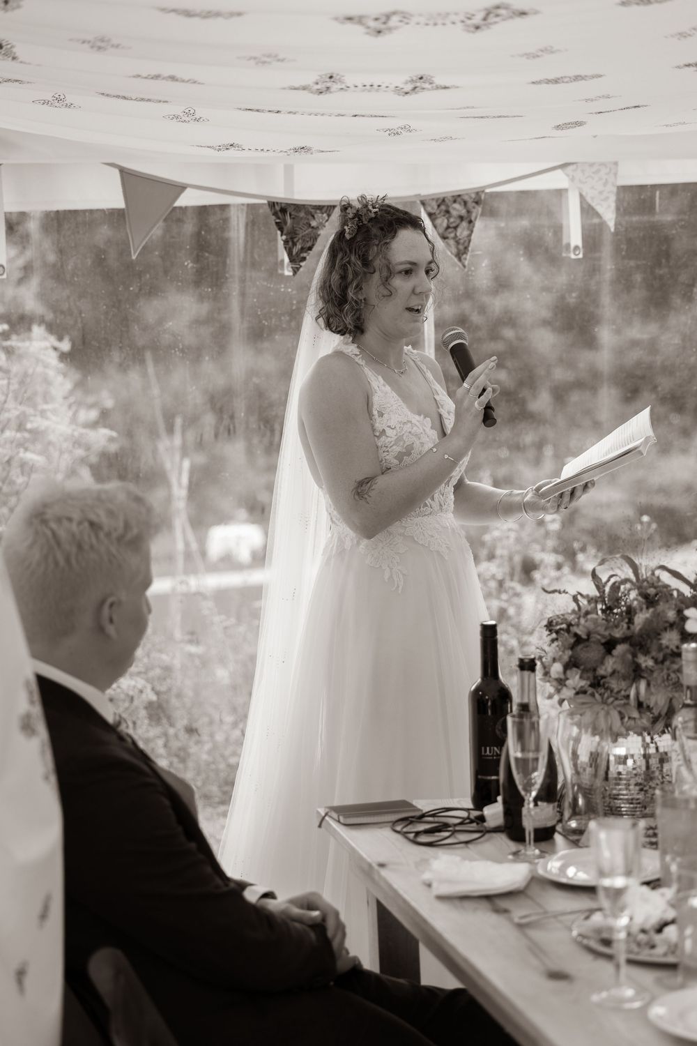 Black and white photo of a bride reading her wedding speech at the elegantly decorated reception table.