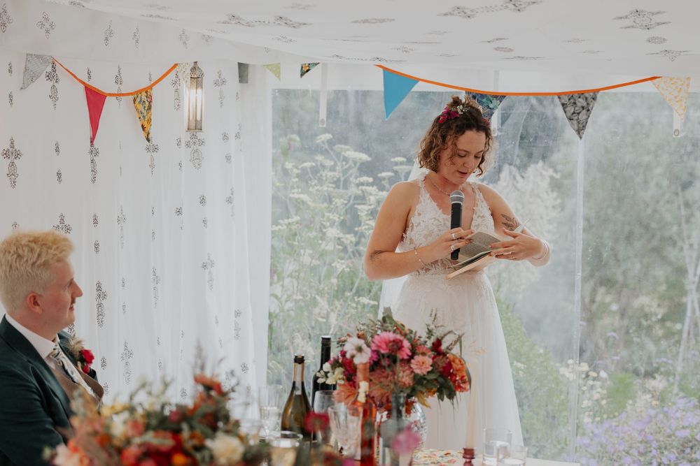 Bride in white dress gives speech at wedding reception in decorated marquee with colorful bunting.