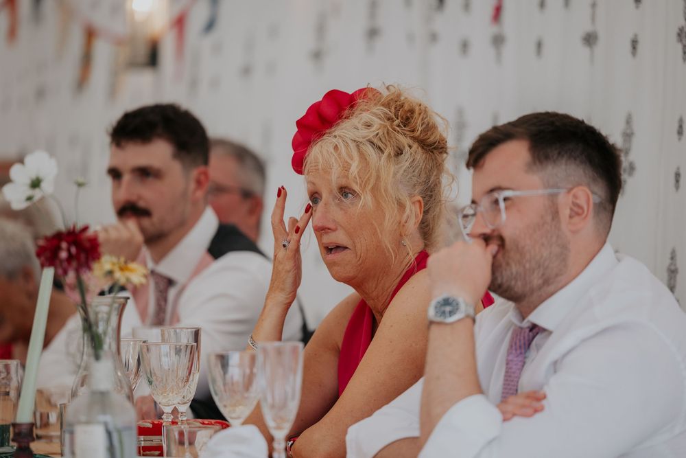 Emotional wedding guest in red dress wipes tear during speeches at elegant reception.