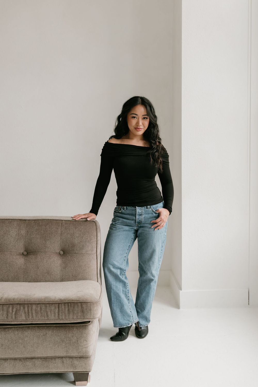 A person in a black long-sleeve top and light wash jeans poses against a white wall with a brown couch.
